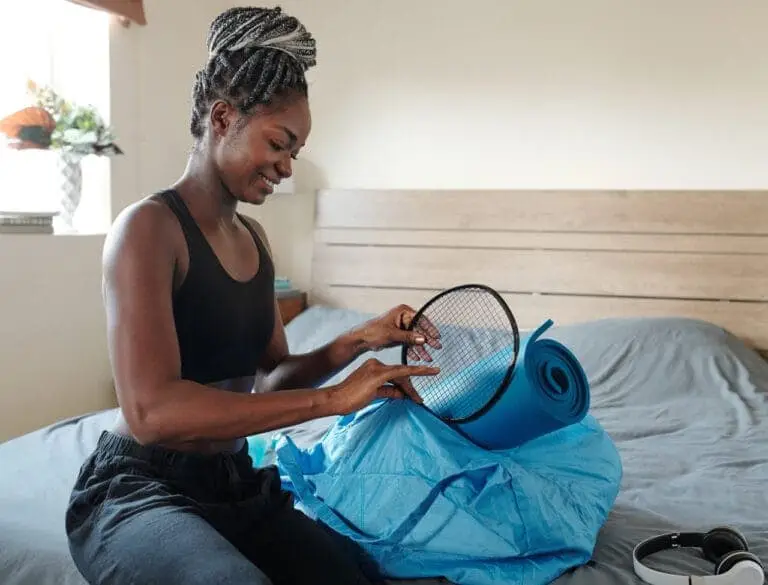 Picture of a Black person packing badminton rackets and a blue yoga mat, on their bed, with wireless headphones