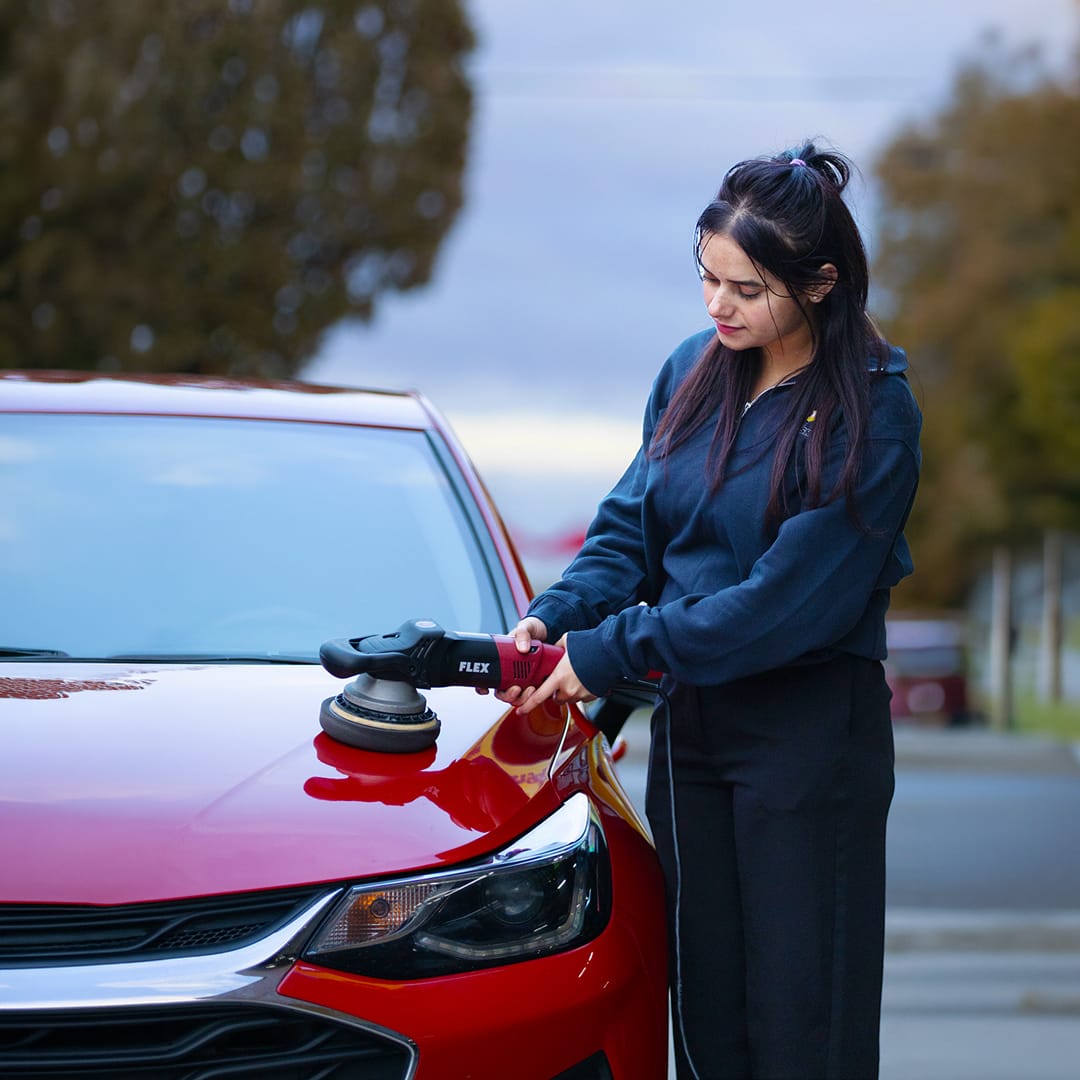 Woman using an electric polisher to buff the hood of a red car outdoors