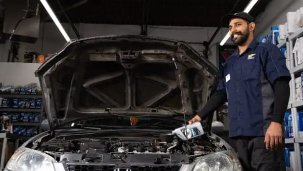 Technician smiling beside a car with open hood, pouring oil into the engine from a bottle