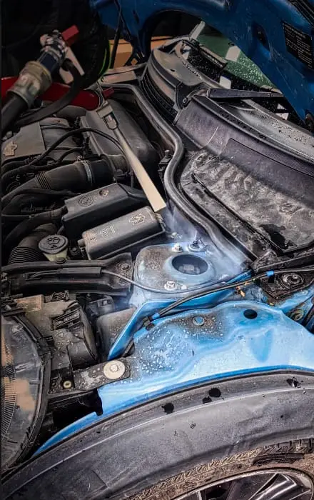 Close-up of a car engine bay with a blue body, showing wet, soapy surfaces during cleaning or detailing