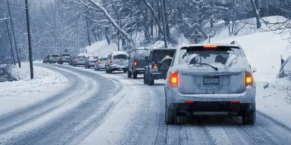 Line of vehicles driving cautiously on a snow-covered road in a winter landscape, with snow coating the cars and trees, illustrating the challenges of winter driving and the importance of keeping car sensors clean and functional.