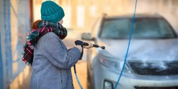 Person bundled in a winter coat, scarf, and beanie using a pressure washer to clean their car at an outdoor car wash, with a frosted vehicle in the background, emphasizing winter vehicle care.