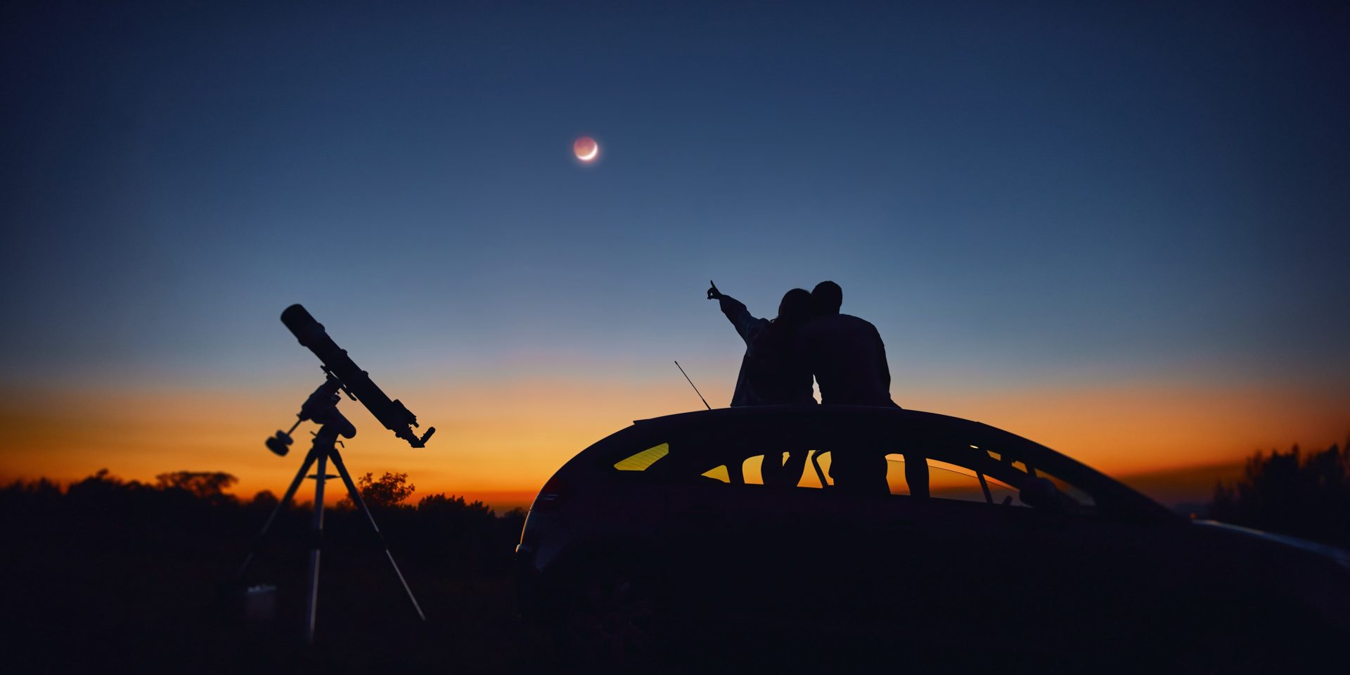 Picture of a couple on the hood of their car, pointing at a solar eclipse, with a telescope in the background