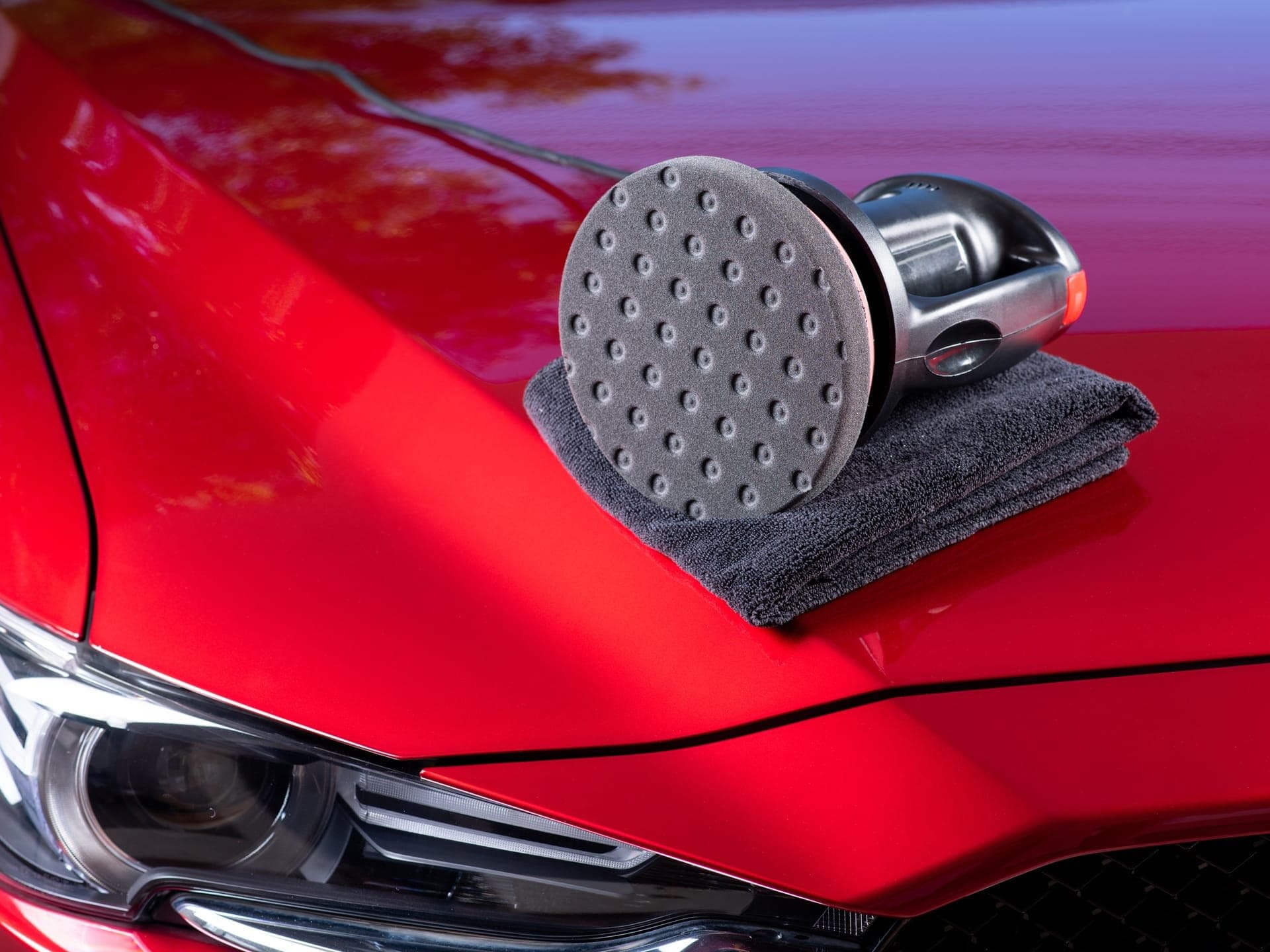 Cordless orbital polisher with foam pad and microfiber towel resting on a glossy red car hood