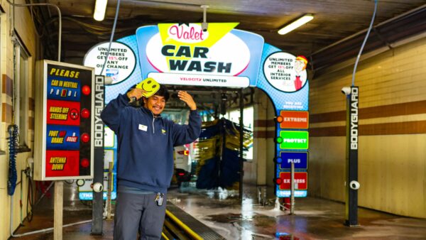 Smiling car wash employee poses with a smiley-face brush at the entrance of Valet Car Wash, with unlimited monthly washes starting at $17.99